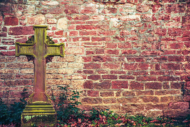 A weathered stone cross covered in vibrant green moss stands prominently against an aged red brick wall. The scene is filled with rustic textures, featuring climbing ivy and scattered autumn leaves, creating a nostalgic and peaceful atmosphere in a historic garden or cemetery.