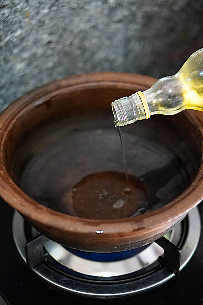 Image of of coconut oil being poured from a bottle into  claypot on stovetop flame, capturing hands-on cooking, food preparation