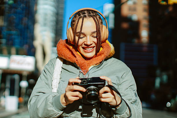 Stylish young female student photographing with a vintage analog camera while wearing large headphones, enjoying music and capturing moments in urban New York environment.