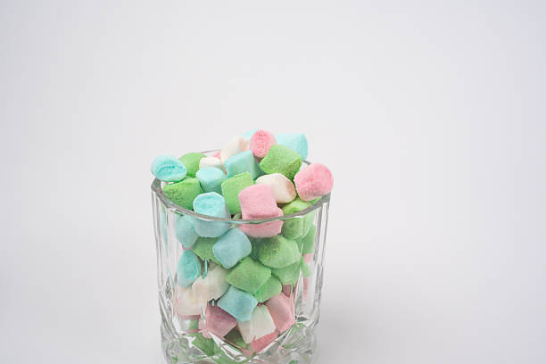 Small pastel colored marshmallows fill a clear glass, presenting a sweet confectionery display. The candies are light green, pink, blue, and white on a clean white background