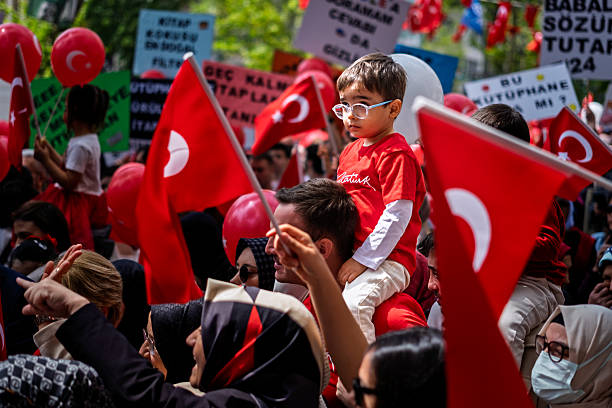 Izmit, Kocaeli, Türkiye - April 23, 2025: A young boy wearing glasses and an Ataturk signature t-shirt sits on his father's shoulders during the National Sovereignty and Children's Day celebrations. The crowd is filled with Turkish flags, red balloons, and protest signs advocating for libraries and reading. The scene captured in the city center reflects the patriotic and educational spirit of the national holiday in Turkey.