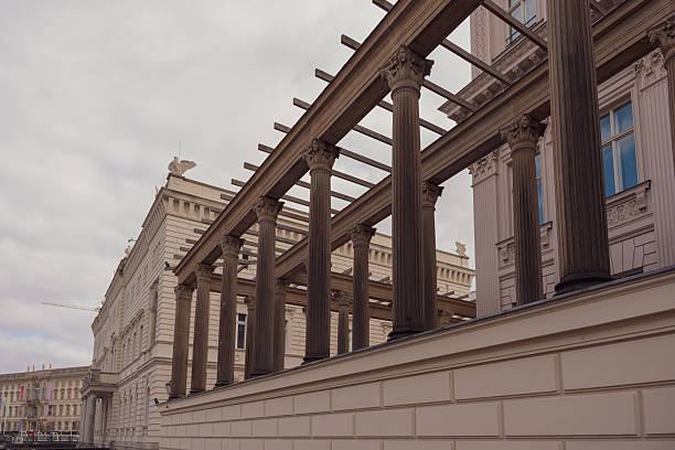 Ancient columns stand tall under overcast sky, framing historic building. Stone pillars show weathered texture and classical design. Urban scene blends architecture with muted tones