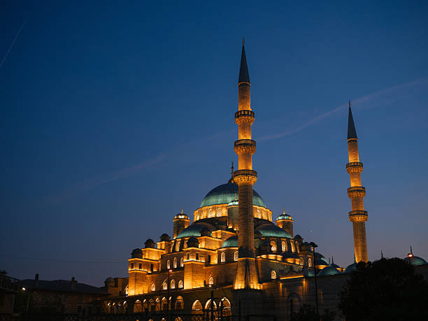 The Yeni Mosque in Istanbul is illuminated with golden lights against a deep blue twilight sky, Turkey