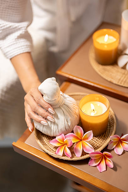 Close up of woman hands holding herbal compress ball with candles and flowers in spa setting. Aromatherapy and wellness concept with warm lighting, relaxation mood, and traditional body treatment in cozy environment.