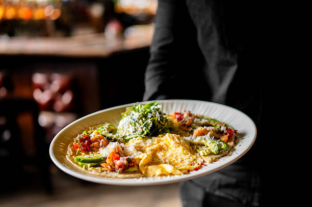 A waiter's hands holding a white plate of Mexican breakfast tacos with scrambled eggs, avocado, salsa, and fresh greens in a dimly lit restaurant.