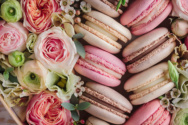 A delightful close-up view of a pastel-colored assortment of macarons, nestled amongst a vibrant bouquet of blooming roses, ranunculus, and delicate white flowers, creating a sweet and elegant visual