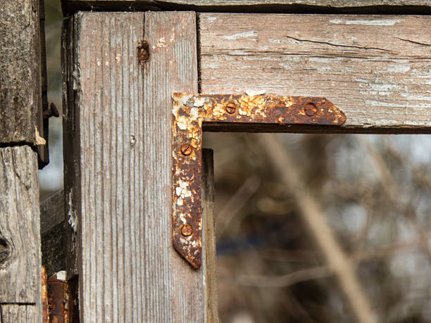 Close-up of a corroded L-shaped metal bracket attached to aging grey wood. The image highlights the detailed texture of rust, peeling white paint, and wood grain, conveying a sense of decay, history, and rustic industrial style. Natural daylight.