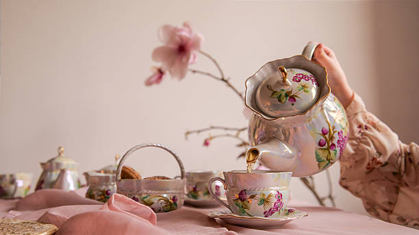 A delicate hand pours tea from a beautifully decorated teapot into a matching teacup, surrounded by other pieces of the set and a soft pink tablecloth