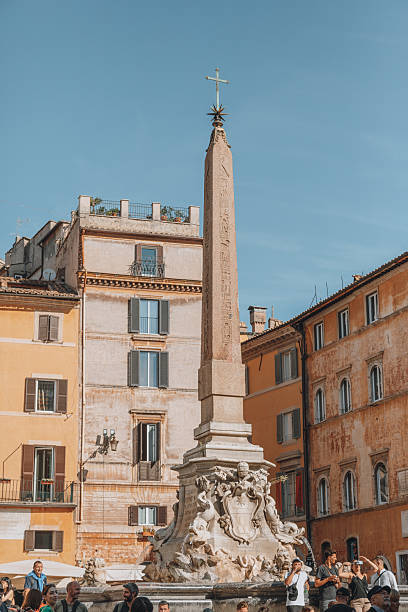 Rome, Italy - View of the Obelisco del Pantheon rising above the Fontana del Pantheon in Piazza della Rotonda. The ancient Egyptian obelisk, crowned with a cross, stands on an ornate Baroque fountain adorned with sculptural figures, surrounded by historic pastel buildings and lively crowds. The scene captures the blend of ancient, Renaissance, and Baroque elements that define the architectural richness of Rome