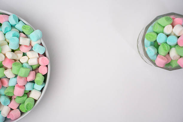 Small pastel marshmallows fill a silver bowl on the left and a clear glass jar on the right. The sweet candies are pink, green, light blue, and white, arranged on a bright white studio surface