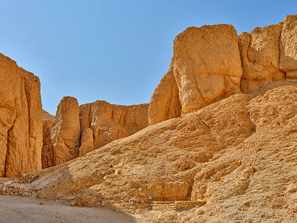 Egyptian desert canyon walls rise in dramatic orange hues beneath a bright blue sky. The rugged rock formations and sandy slopes create a stark, timeless landscape perfect for travel, geology, exploration, and documentary storytelling in stock photo catalogs.