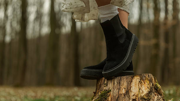 A person's legs are shown from the knees down, wearing casual boots and a floral dress while standing on a mossy tree stump outdoors