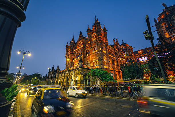 Iconic Mumbai landmark bathed in warm evening light, showcasing ornate Victorian-Gothic architecture, busy street traffic, taxis and commuters, and lively urban atmosphere during blue hour in the city.