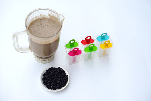 Homemade black bean smoothie in blender with colorful popsicle molds on white background, healthy dessert preparation concept with natural ingredients for refreshing frozen treats.