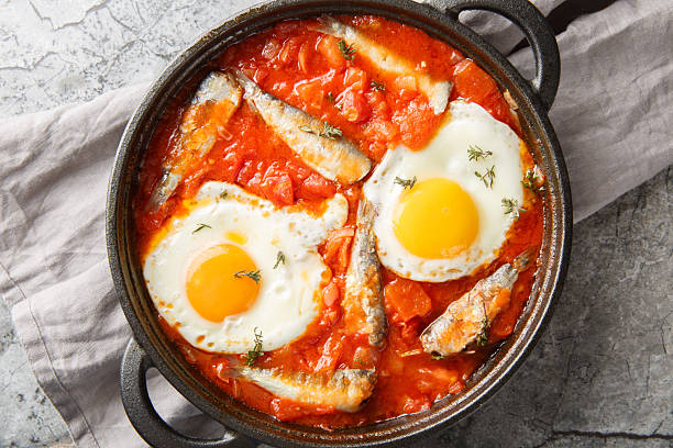 Delicious fried eggs with sardines in vegetable tomato sauce close-up in a frying pan. Horizontal top view