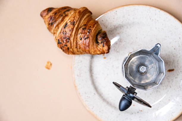 Open and empty Italian coffee maker with a french buttery pastry on a plate on beige background