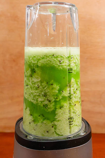 Blender cup with green kiwi mixture and foam on warm wooden background.