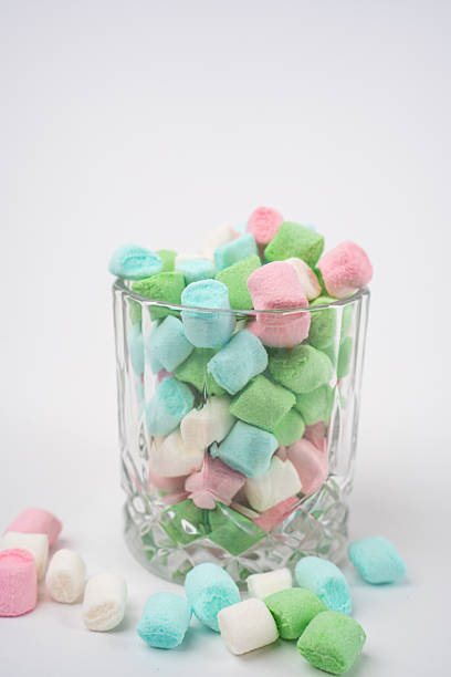A clear glass tumbler full of pastel pink, blue, green, and white mini marshmallows. Scattered marshmallows lie on the clean white surface nearby