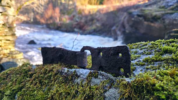 Close-up of heavily corroded antique iron hardware resting on a moss-covered granite rock beside rushing rapids, remnant of a historic watermill in Østfold, Norway.