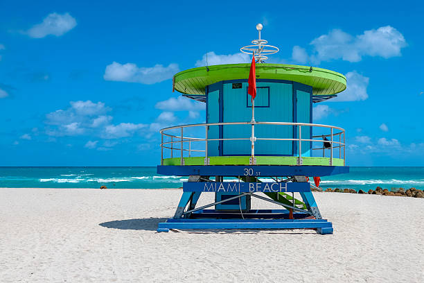 Colorful lifeguard tower standing on the white sand of Miami Beach, Florida, with turquoise ocean, blue sky and soft clouds in the background.