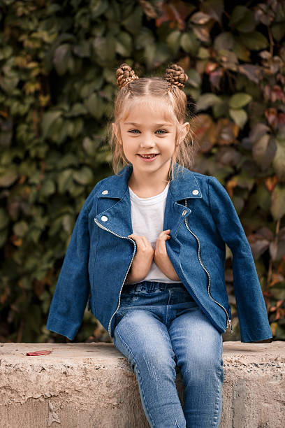 A beautiful stylish blonde girl in a blue jacket sits on a concrete block. Girl 7 years old with a smile on a background of leaves