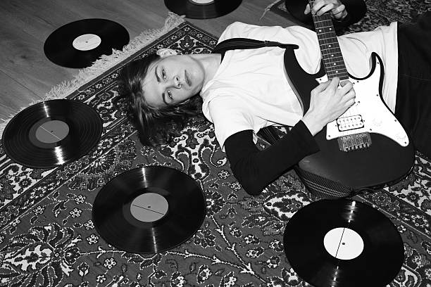 A young man with an electric guitar lies on a carpet surrounded by records