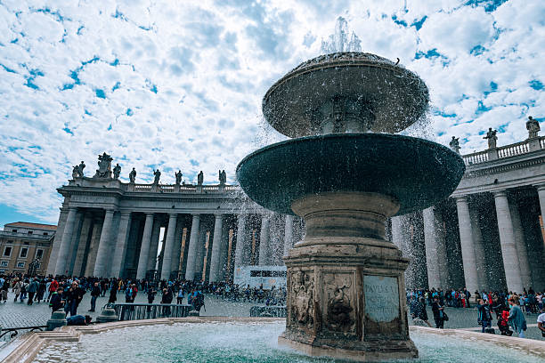 Rome, Italy - May 3, 2019: Water sprays from a fountain in St Peter’s Square as crowds gather near the colonnade and basilica in Vatican City.