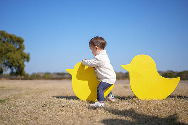 Little child pushes giant yellow bird cutout. Bright blue sky, dry grass, distant tree. Child smiles, full of joy and curiosity. Outdoor playtime, springtime, family fun. Perfect for parenting