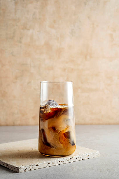 Freshly poured milk creates beautiful swirls in a tall glass of iced coffee. The drink is presented on a minimalist stone coaster against a neutral background.