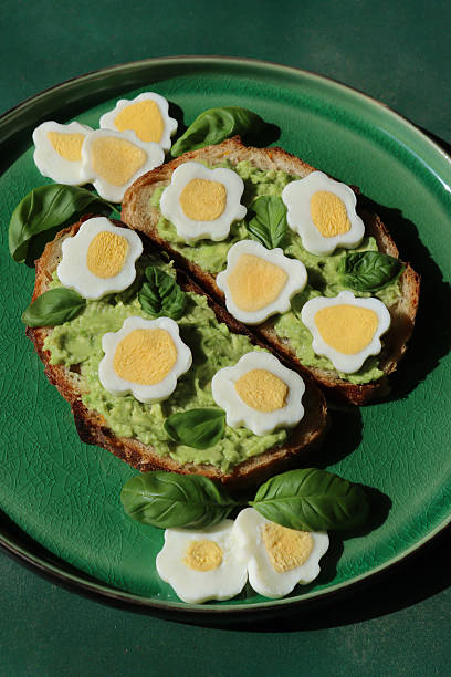 Close-up image of Easter breakfast, emerald green, crackle glazed plate containing flower-shaped, sliced hard-boiled eggs with bright yellow yolks, served on sourdough toast topped with mashed avocado, basil leaf herb garnish, green tile background