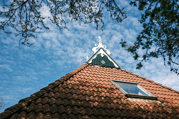 Leeuwarden, Friesland, The Netherlands. April 4th, 2026: Upward view of a traditional Dutch building with a red tiled roof and decorative white wooden gable, framed by tree branches and set against a bright blue sky with scattered clouds.