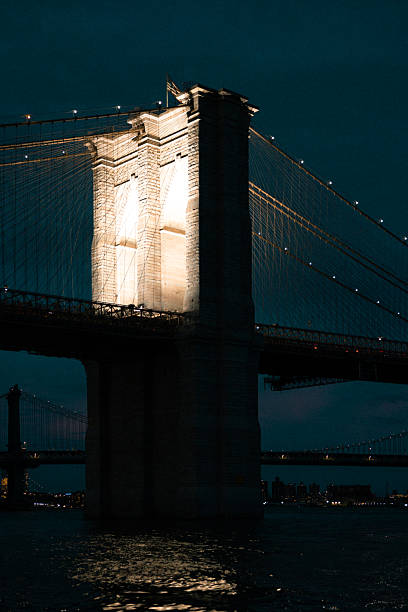 Brooklyn Bridge's stone towers are illuminated against a dark night sky, spanning the water with its iconic cables and lights reflecting on the river below