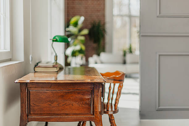 Vintage wooden desk with green lamp and books near window in bright modern interior, cozy workspace concept.