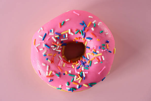 Close-up image of glazed ring doughnut, pink fondant iced ring deep-fried dough dessert decorated with multicoloured hundreds and thousands sugar sprinkles, pink background, elevated view