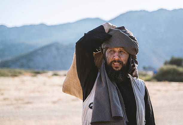 Portrait of a refugee man carrying a sack over his shoulder in a dry arid landscape with mountains in the background