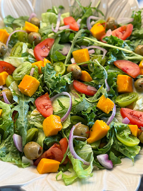 Full frame image of white, sunflower design salad bowl of feta cheese mixed salad with lettuce and rocket leaves, red onion slices, mango fruit cubes, tomato quarters, green cherry tomato halves, green olives, elevated view, healthy eating concept