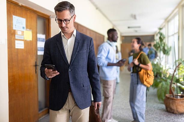 Professor walking trough university hallway using mobile phone while students pass by stock photo