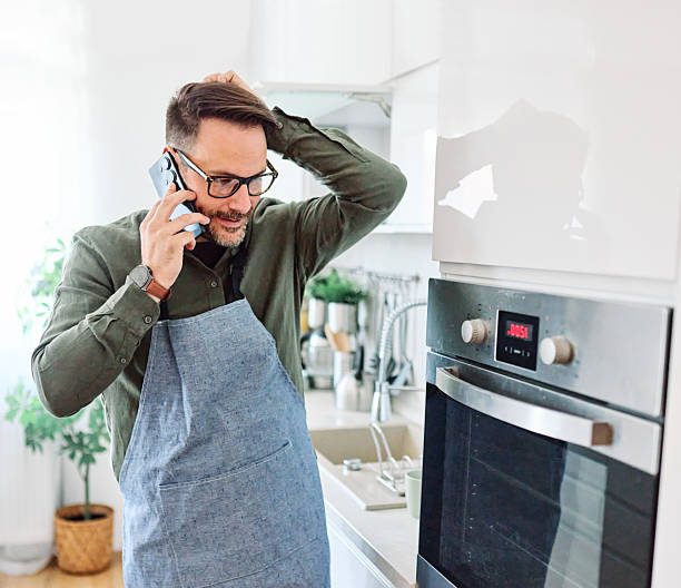 man kitchen smartphone apron browsing phone mobile cooking technology hobby home conversation stress multitasking support help talking busy problem A bearded man wearing glasses and a blue apron talks on his phone in a bright modern kitchen, appearing concerned while checking the oven. Natural light, home appliances and plants set a domestic scene as he juggles a phone call and cooking tasks, conveying stress, multitasking and everyday life. What to Check Before Calling an Appliance Repair Professional stock pictures, royalty-free photos & images