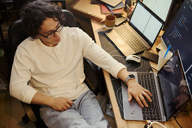 Man Coding on Laptop at Home Office Desk with Multiple Screens stock photo