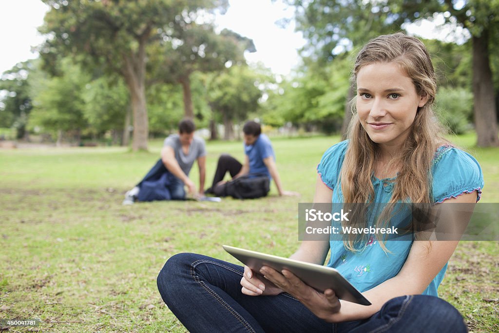 Blonde student using a tactile tablet while sitting Blonde student using a tactile tablet while sitting in a park with friends studying in background 20-29 Years Stock Photo