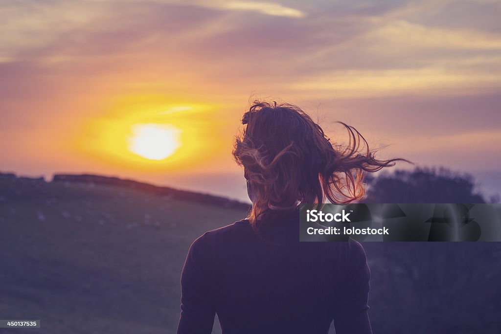 Young woman admiring the sunset over fields Rear view of young woman admiring the sunset over a field with her hair blowing in the wind Sunset Stock Photo