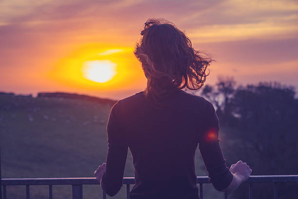 Woman admiring sunset from her balcony Rear view of young woman admiring the sunset over a field from her balcony looking through window stock pictures, royalty-free photos & images