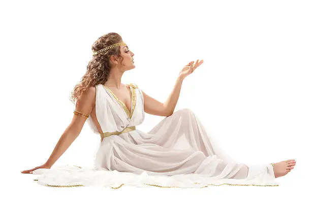 The Beautiful Young Woman Sitting on the Floor Wearing White and Gold Greek Costume, Raising Her Arm on the White Background . 