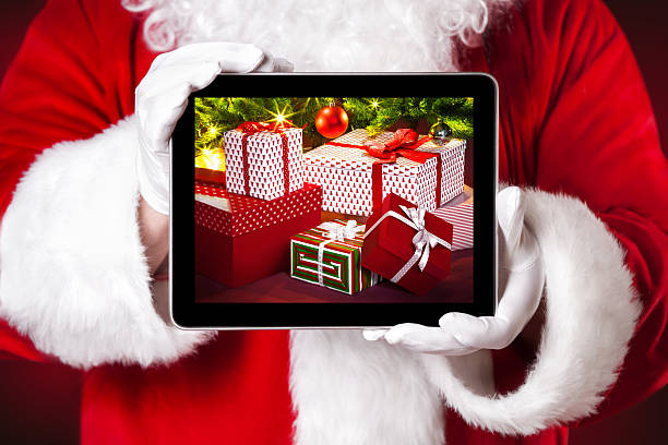 Santa holding a tablet which shows Christmas presents stock photo
