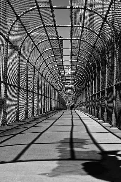 A DSLR photo of a fenced-in pedestrian overpass of a city. The photo is in black and white. The curving fence casts straight shadows along the sidewalk