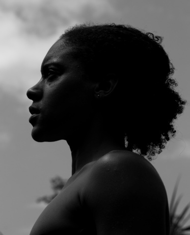 Low angle moody black and white portrait of a beautiful young African woman standing outdoors staring into distance, profile view