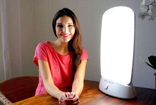 Beautiful young lady sitting in front of her full-spectrum light. Light therapy which strikes the retina of the eyes is used to treat circadian rhythm disorders such as delayed sleep phase disorder and can also be used to treat seasonal affective disorder as well as to fight winter depression.