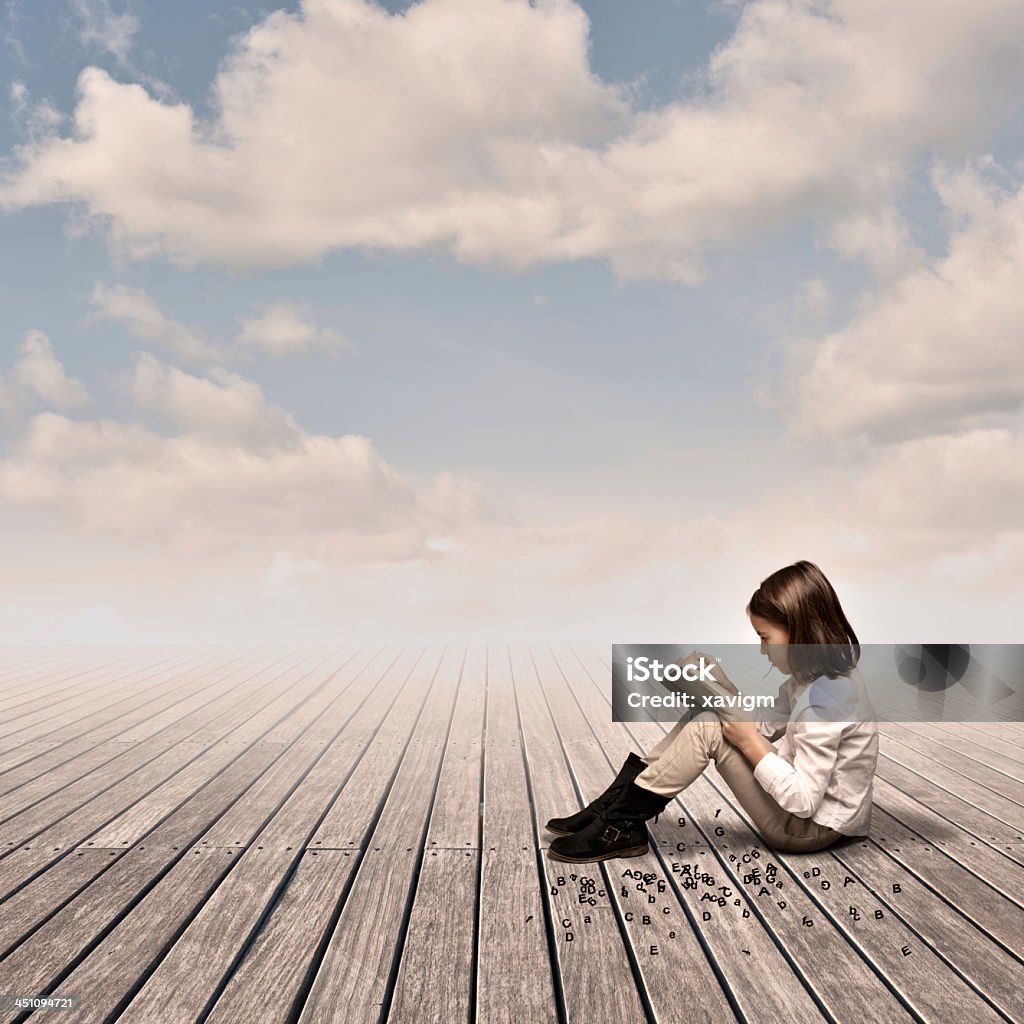 Young girl reading on a wooden surface under a cloudy sky little girl reading a book on a wharf Girls Stock Photo
