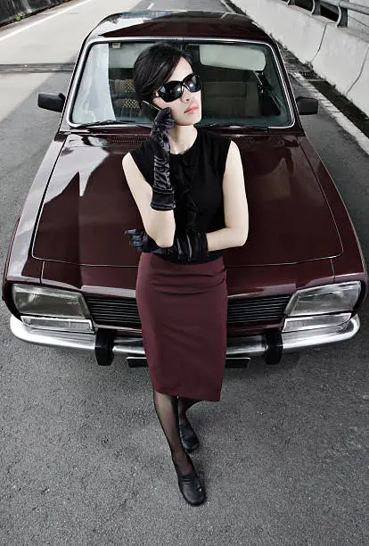 An Asian woman in 60's wardrobe in front of a vintage Peugeot 504 car using her cell phone.