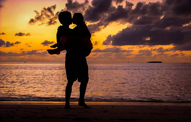 Man Holding a Woman Kissing in Front of the Sunset Silhouette of a man holding a woman in his arms, kissing on the beach in front of a red sunset with a distant island in the background. Taken on the island of Meeru in the Maldives. meeru island stock pictures, royalty-free photos & images